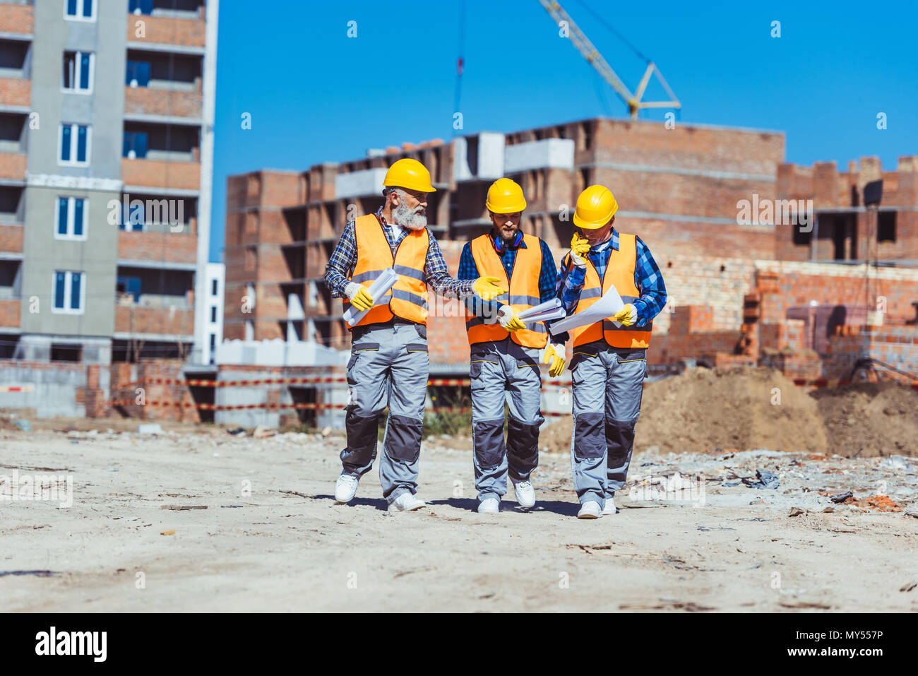 Drei Arbeiter in Warnwesten und hardhats zusammen gehen über Baustelle und diskutierten Pläne Stockfoto