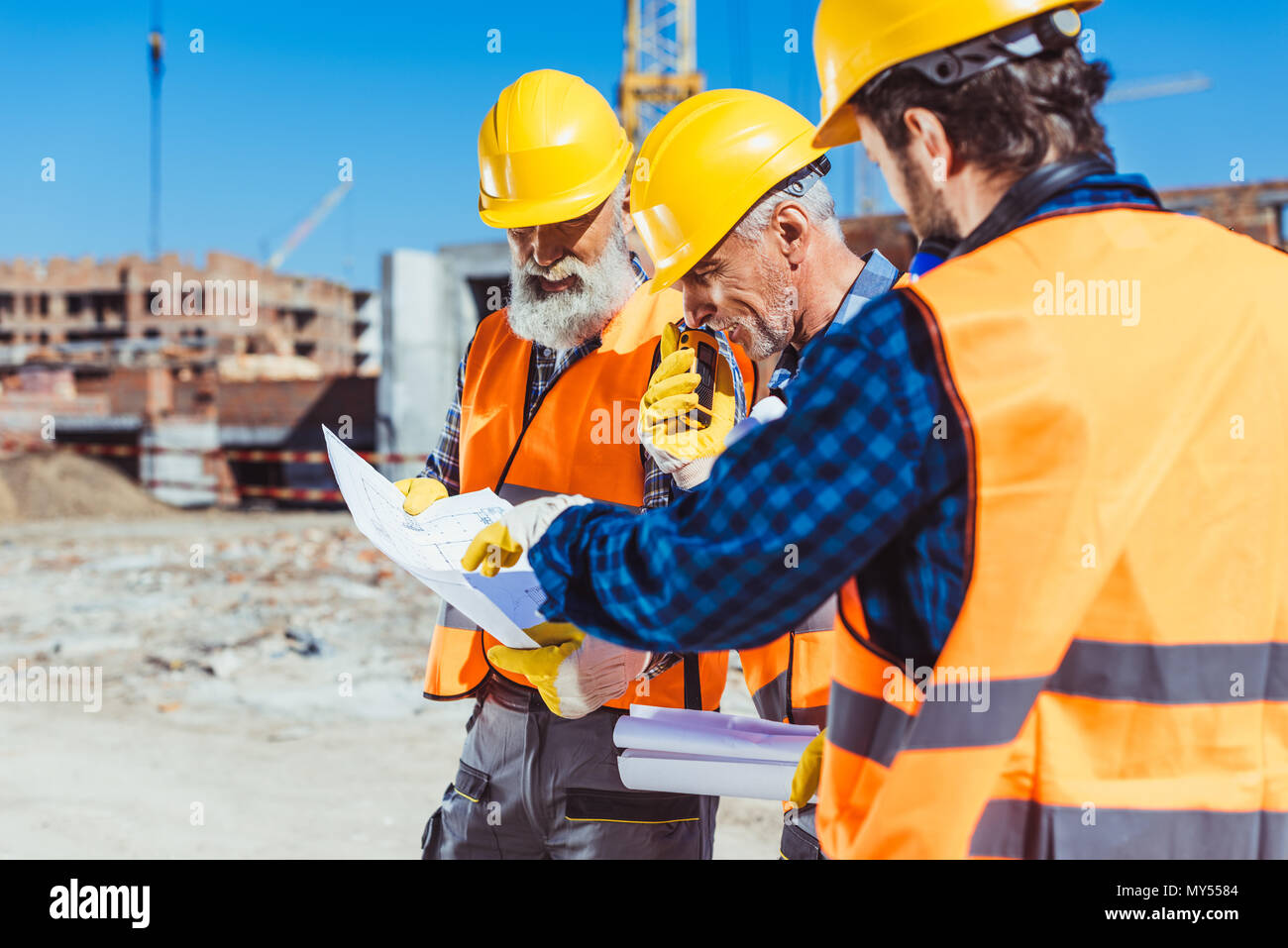 Drei Arbeiter in Warnwesten und hardhats an der Baustelle stehen und Prüfung der Pläne Stockfoto