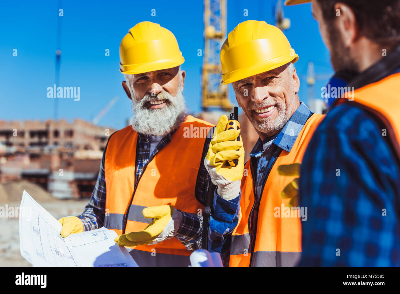 Zwei Arbeiter in Warnwesten und hardhats Prüfung Baupläne und Sprechen auf tragbaren Radio auf der Baustelle Stockfoto