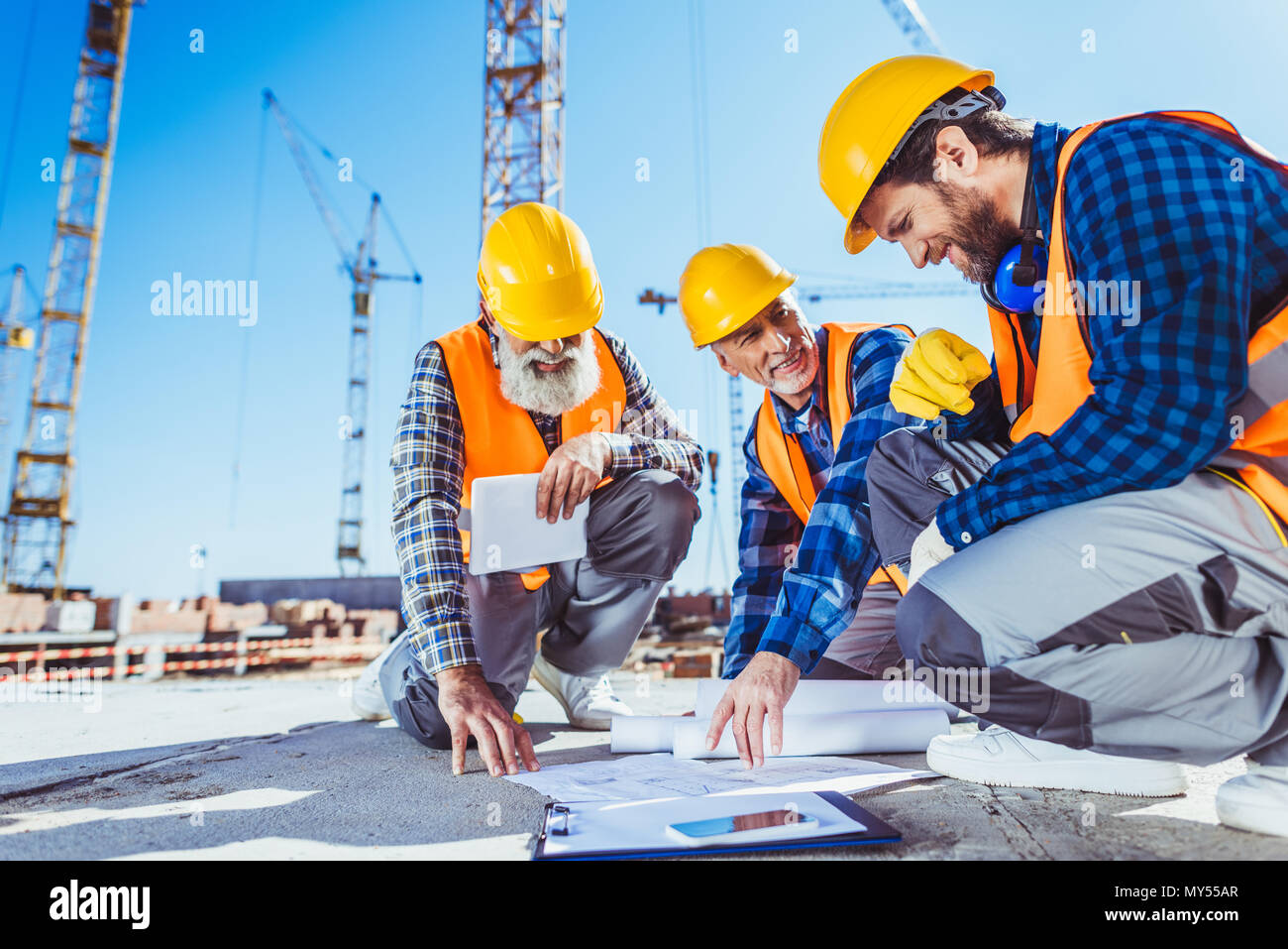 Drei Bauarbeiter sitzen auf Beton auf der Baustelle, diskutieren Baupläne Stockfoto