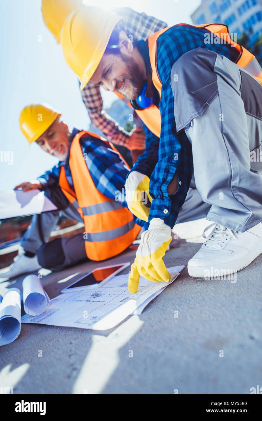 Lächelnd Bauarbeiter in hardhats sitzen auf Beton bei Bau, Prüfung der Pläne Stockfoto