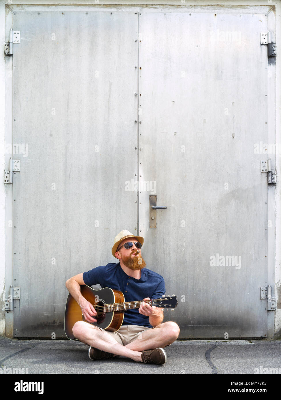 Foto eines jungen männlichen Erwachsenen das Rauchen einer Zigarre und eine akustische Gitarre spielen auf der Straße. Stockfoto