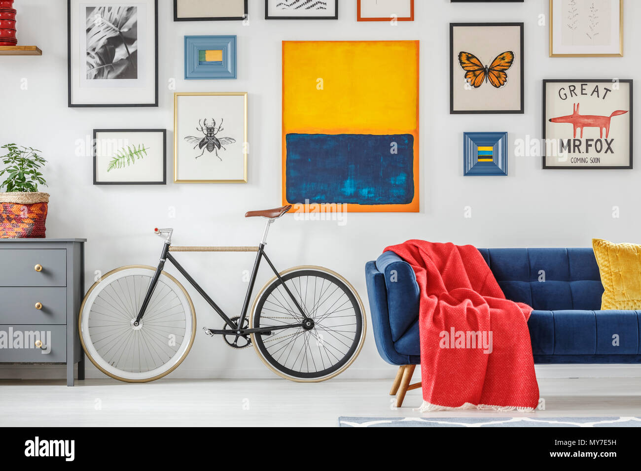 Leuchtend rote Decke auf ein navy blau Sofa neben einem Vintage Fahrrad gegen weiße Wand mit einer Galerie von Plakaten im Wohnzimmer. Real Stockfoto