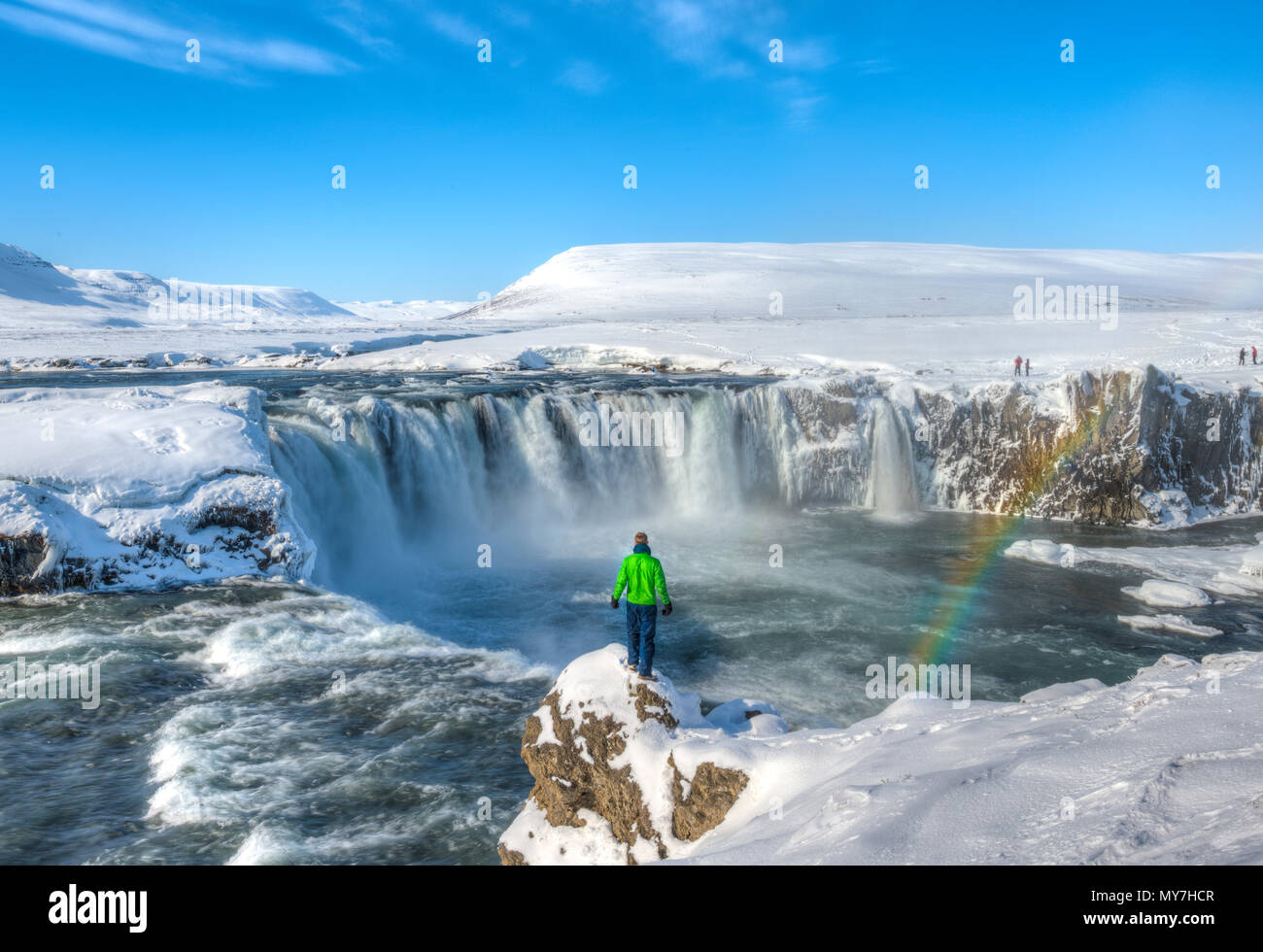 Junge Mann, der auf einem Felsen, Wasserfall mit Regenbogen, Góðafoss, Godafoss im Winter mit Schnee und Eis, Norðurland djupivogur Stockfoto