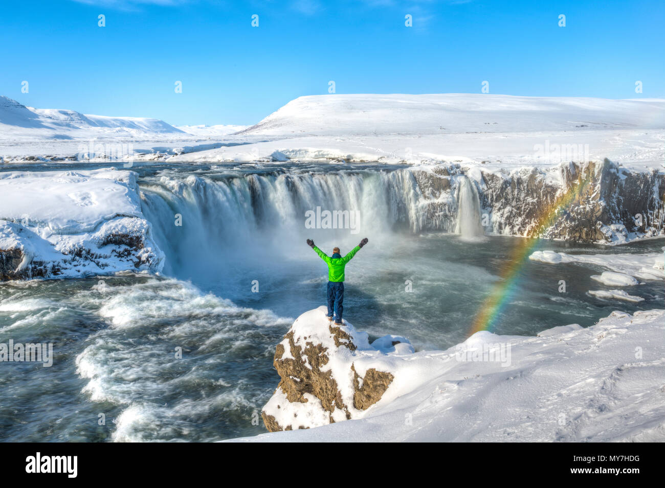 Junger Mann, auf einem Stein und streckte seine Arme in die Luft, Wasserfall mit Regenbogen, Góðafoss, Godafoss im Winter mit Stockfoto
