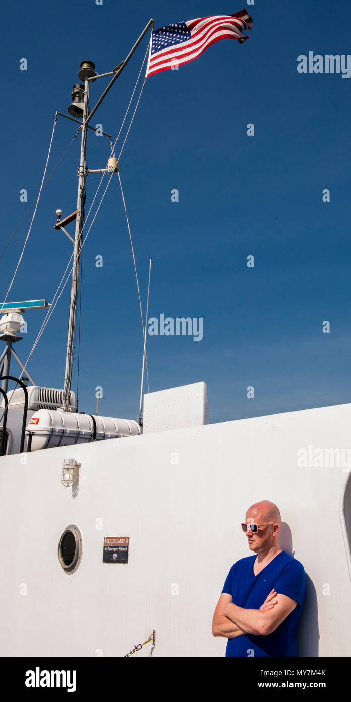 Mann trägt ein blaues T-Shirt, gegen ein helles weißes boot, blauer Himmel lehnend, mit der amerikanischen Flagge im Hintergrund, New York City, USA Stockfoto