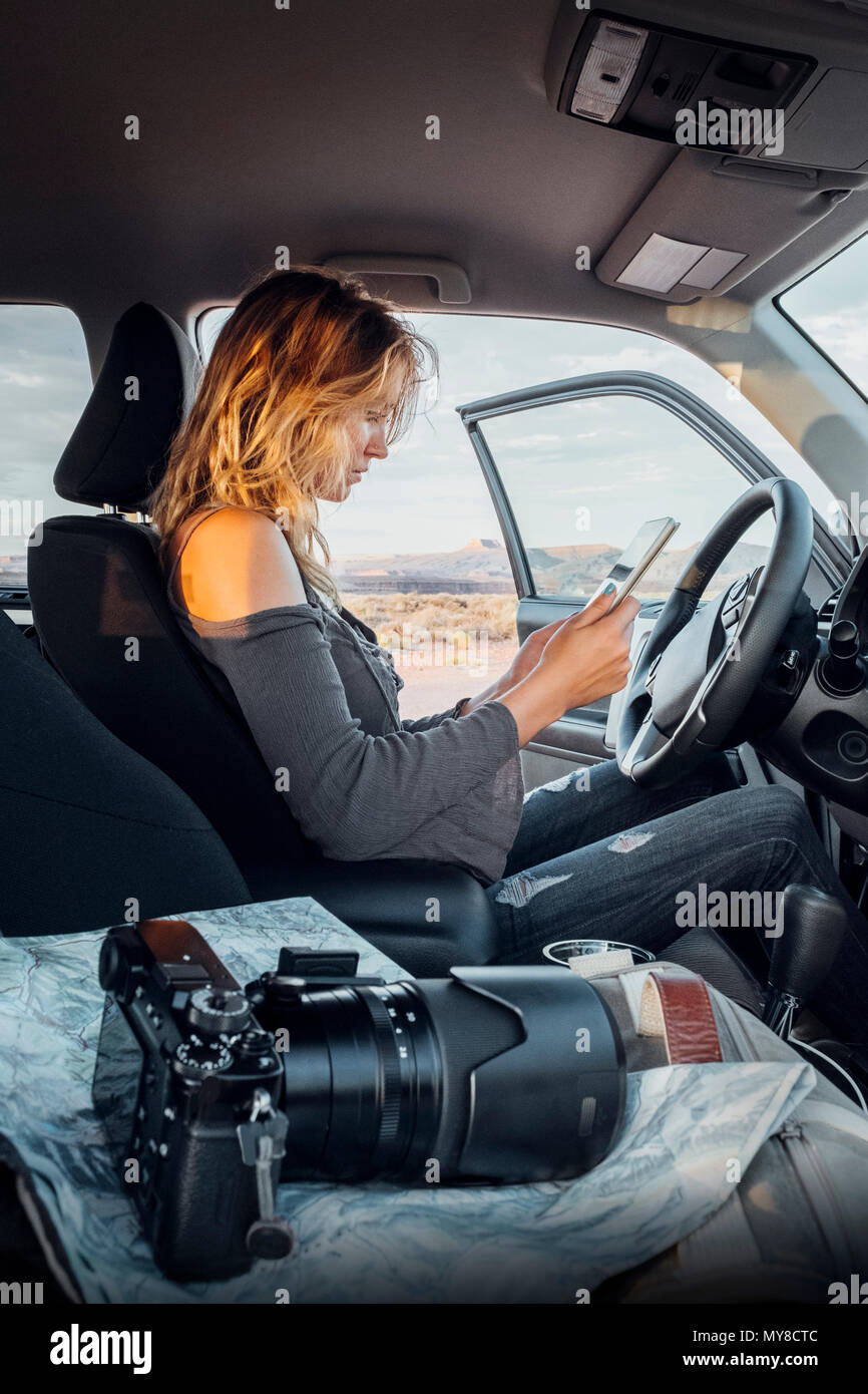 Junge Frau im Fahrzeug sitzen, bei digital Tablet suchen, SLR-Kamera auf Beifahrersitz, Mexican Hat, Utah, USA Stockfoto
