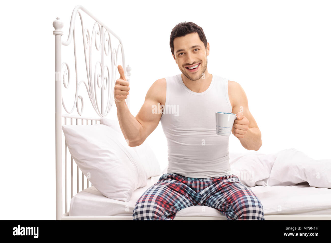 Lächelnden jungen Mann mit einer Schale sitzt auf einem Bett und ein "Daumen hoch" Geste auf weißem Hintergrund Stockfoto