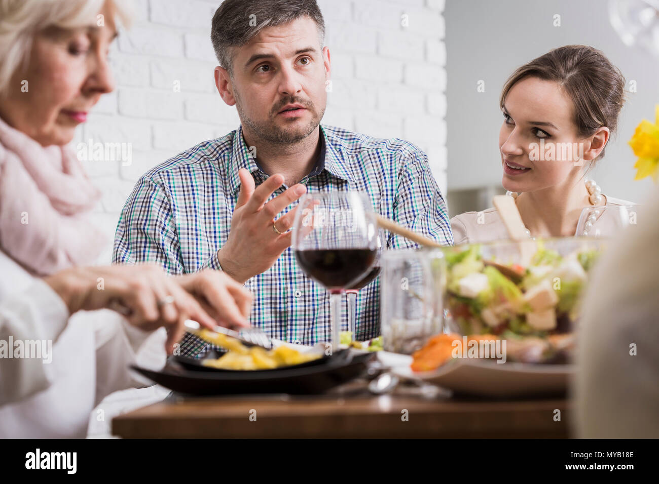Junger Mann und zwei Frauen sitzen neben Tisch beim Abendessen mit der Familie Stockfoto