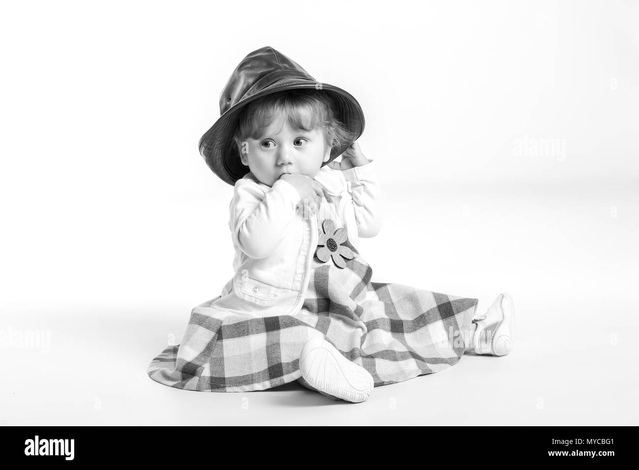 Elegante Happy Baby girl 1 Jahr alt sitzen auf dem Studioboden mit schwarzem Leder Hut. Weißer Hintergrund. Konzept Das Glück. Stockfoto