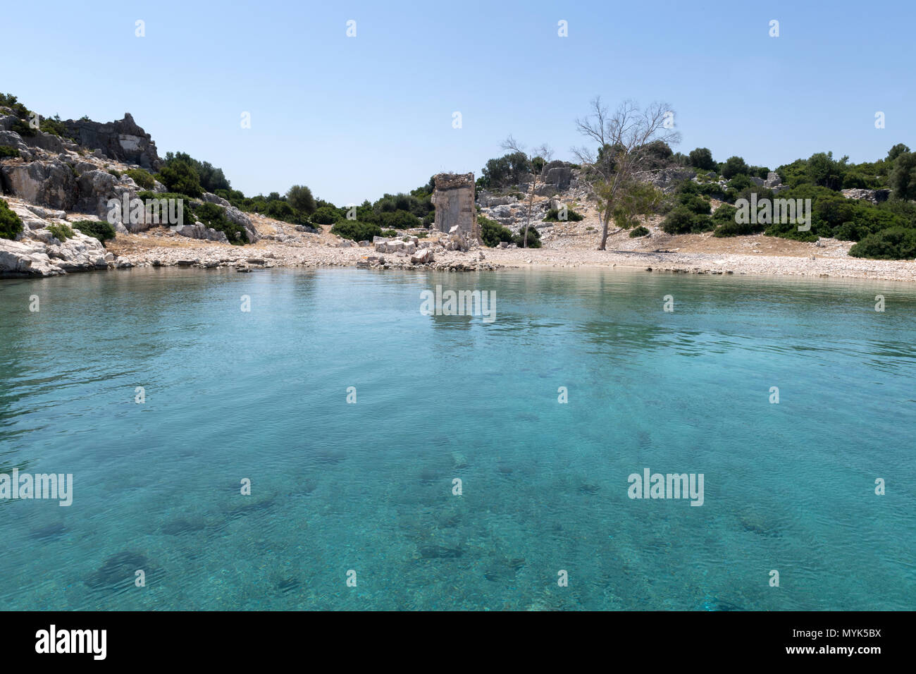 Ein atemberaubender Blick auf eine ruhige Bucht mit türkisfarbenem Wasser und antiken Ruinen in der Ferne, umgeben von felsigem Gelände, Stockfoto