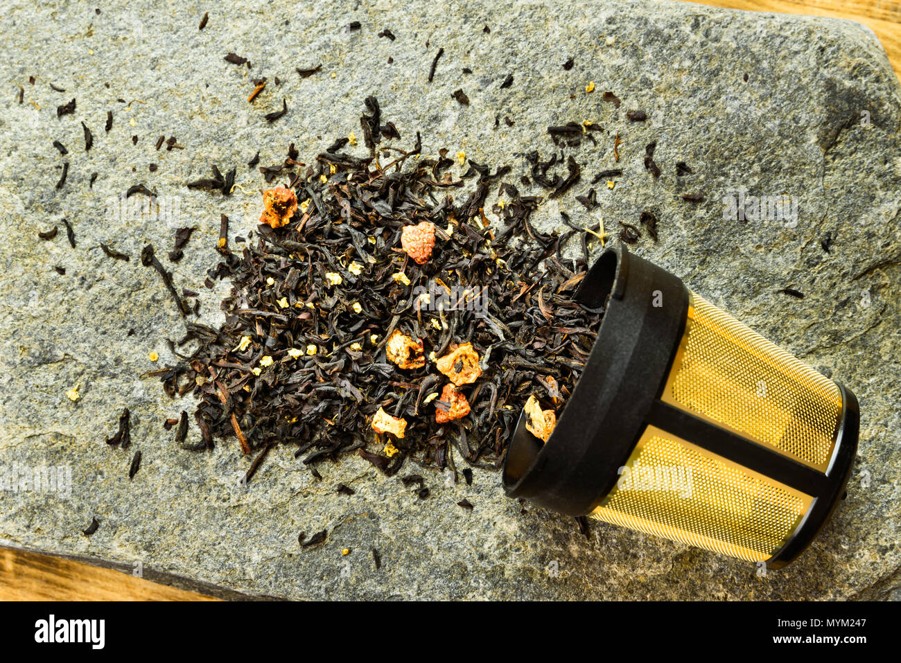 Frucht Aromatisierter schwarzer Tee außerhalb einer vergoldeten Tee-ei auf einer Steinplatte gestreut. Stockfoto