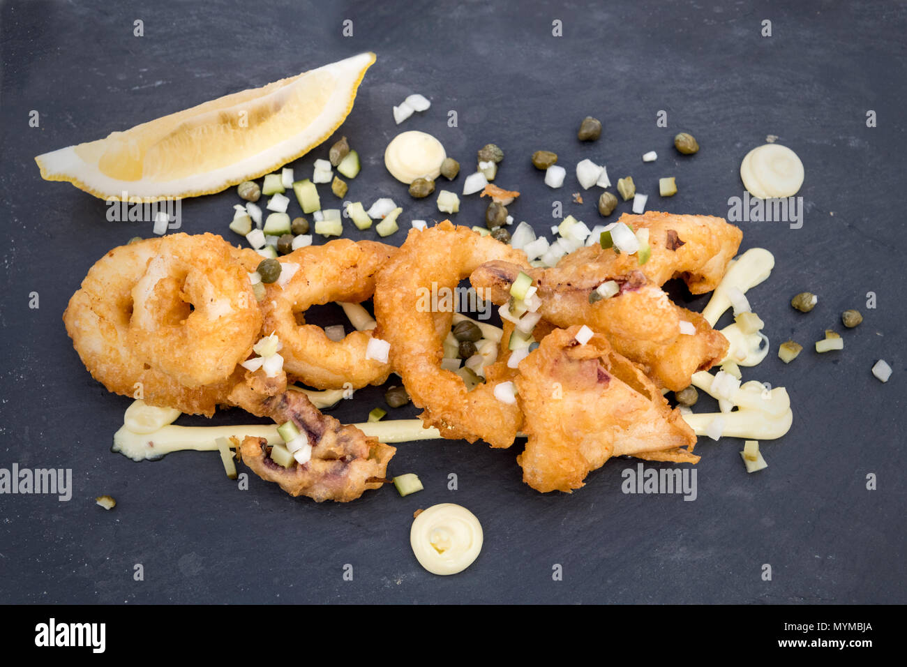 Ein köstlicher Teller mit goldgebratenen Calamari-Ringen, serviert mit einer Scheibe Zitrone, garniert mit fein gehackten Zwiebeln, Kapern, Stockfoto