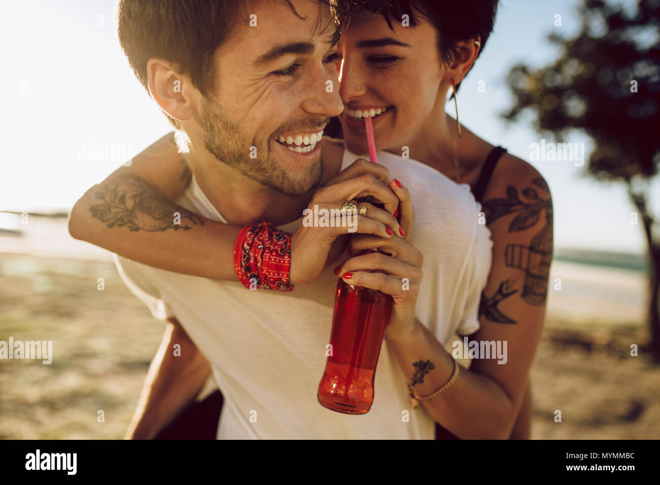 Hübscher junger Kerl huckepack seine Freundin trinken Getränk im Freien. Happy kaukasischen Paar selbst im Freien genießen. Stockfoto
