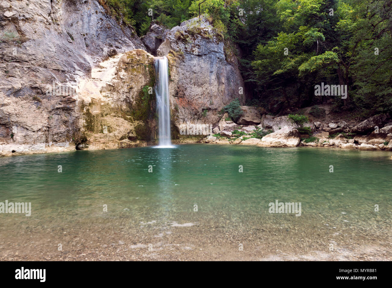 Ein friedlicher Wasserfall, der in einen kristallklaren, smaragdgrünen Pool stürzt, umgeben von felsigen Klippen und üppigem Grün, Stockfoto