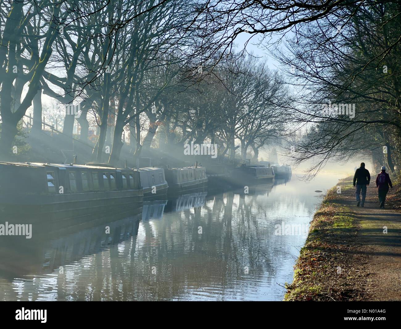 Wetter in Großbritannien: Sonnig und frostig in Adlington. Reflexionen von schmalen Booten und Bäumen auf dem Leeds- und Liverpool-Kanal bei Adlington in Lancashire mit zwei Personen, die auf einem Schleppweg laufen Stockfoto