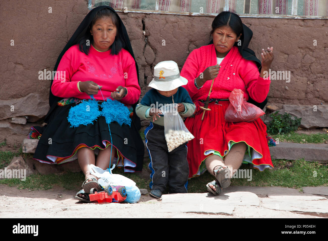 Ein junges Kind seinen Beutel mit Süßigkeiten inspiziert, während seine Mutter und eine andere Frau ihren Nachmittag verbringen Stricken auf Taquile Insel, den Titicacasee. Peru Stockfoto