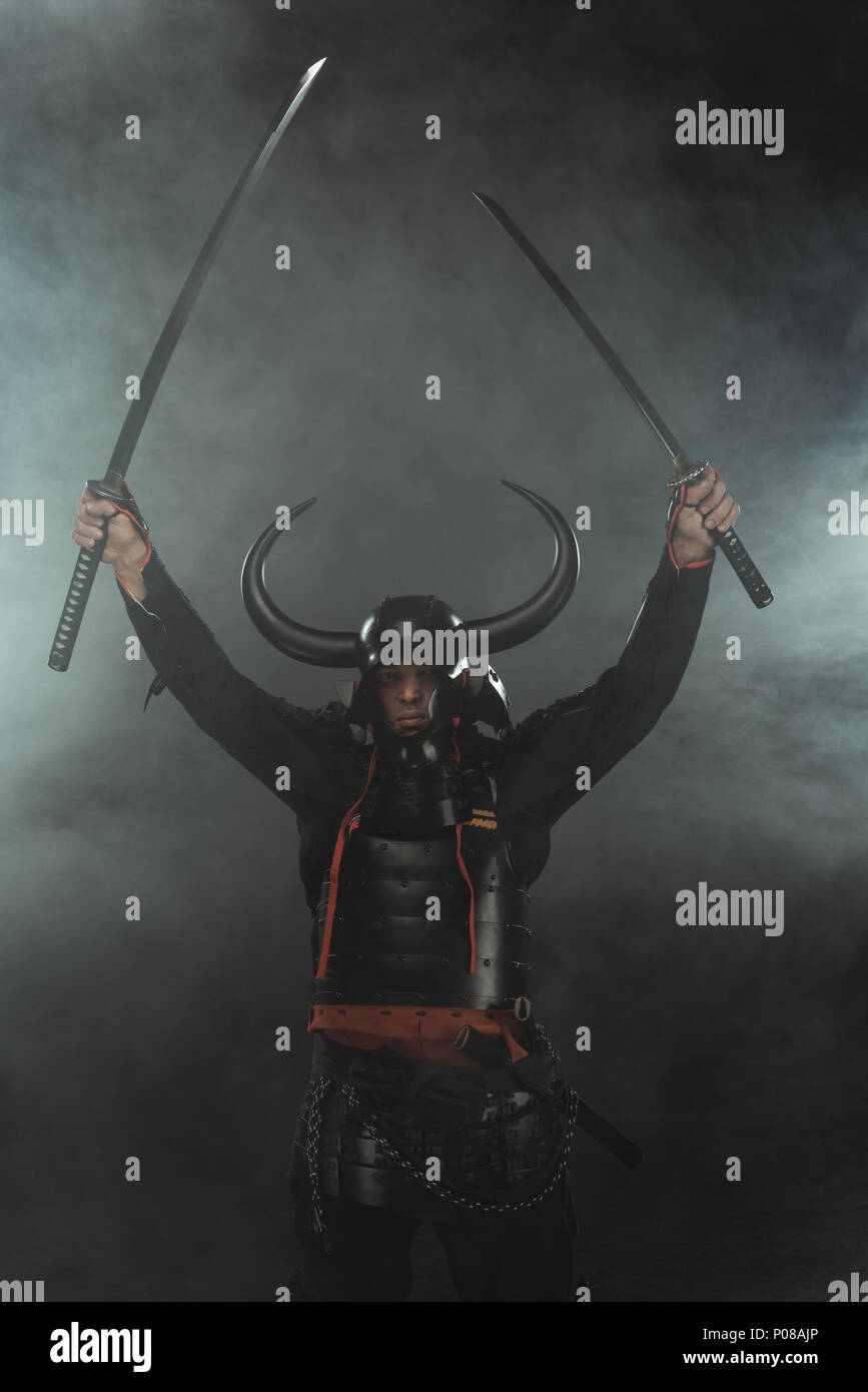 Samurai in Rüstung mit zwei Katana Schwerter auf dunklem Hintergrund mit Rauch Stockfoto