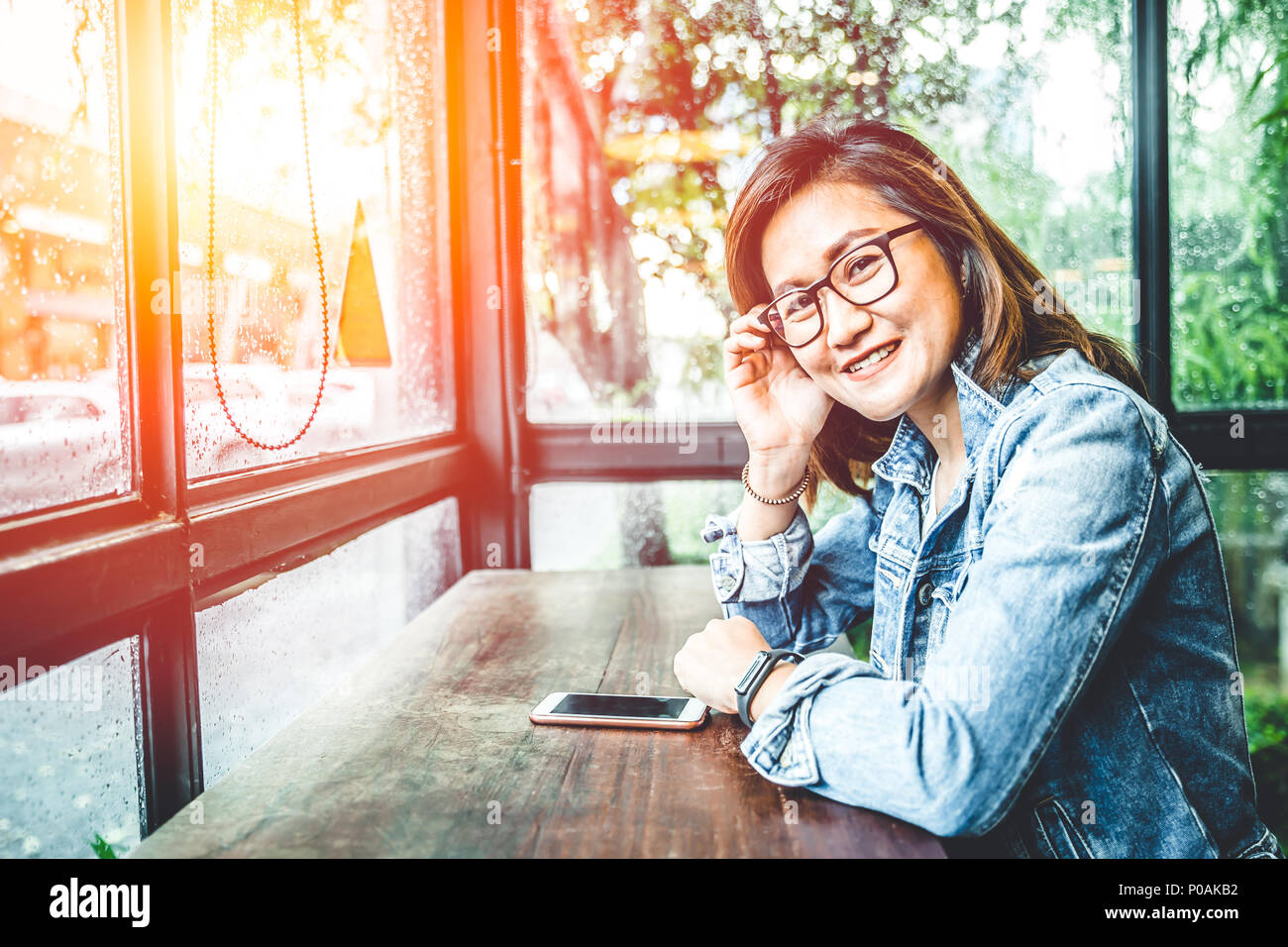 Brille nerd Hipster asiatische Frau sitzen lächeln in Glas windows Cafe mit Smartphone Stockfoto