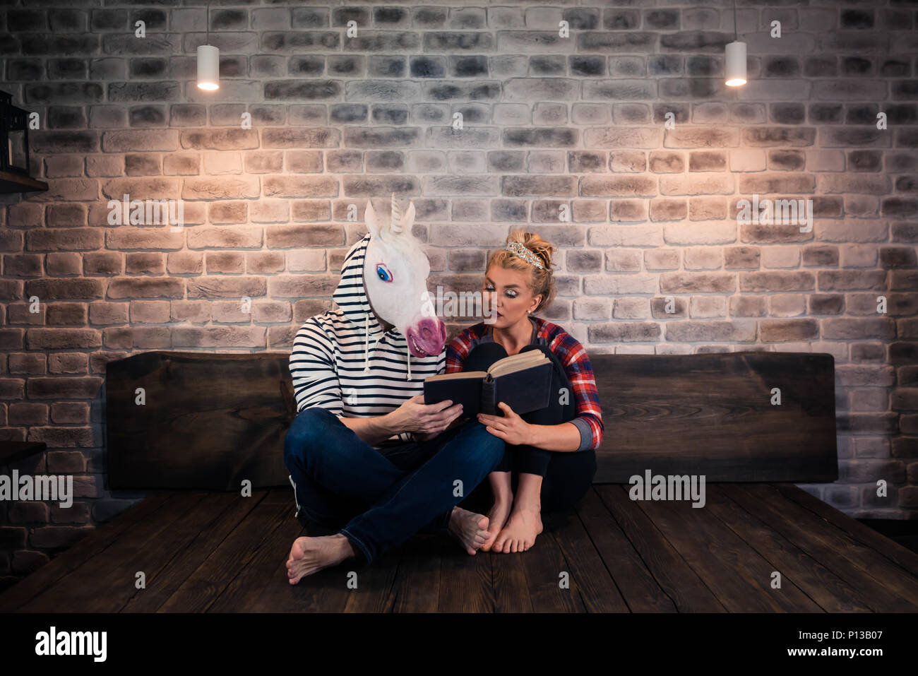 Ungewöhnliche Paar ein Buch lesen in stilvolle Apartment. Aufmerksame Mädchen sitzt auf dem Bett mit lustigen Mann in komischen Maske. Junge Frau mit seltsamen Freund Stockfoto