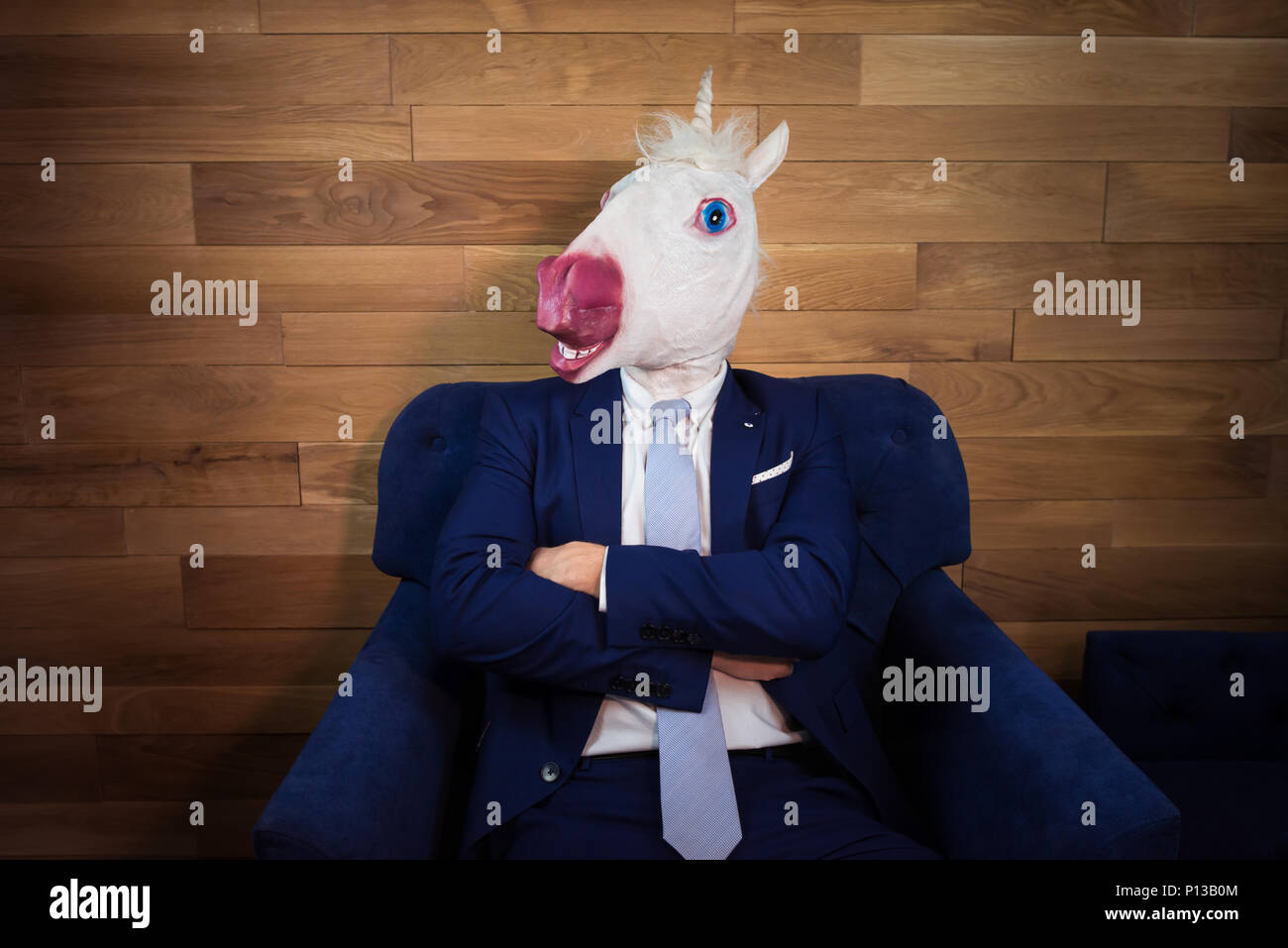 Portrait von ungewöhnlichen Menschen im Home Office. Freaky junge Manager in komischen Maske auf dem Hintergrund der hölzernen Wand. Unicorn in Anzug sitzt auf einem Sessel wie Boss Stockfoto