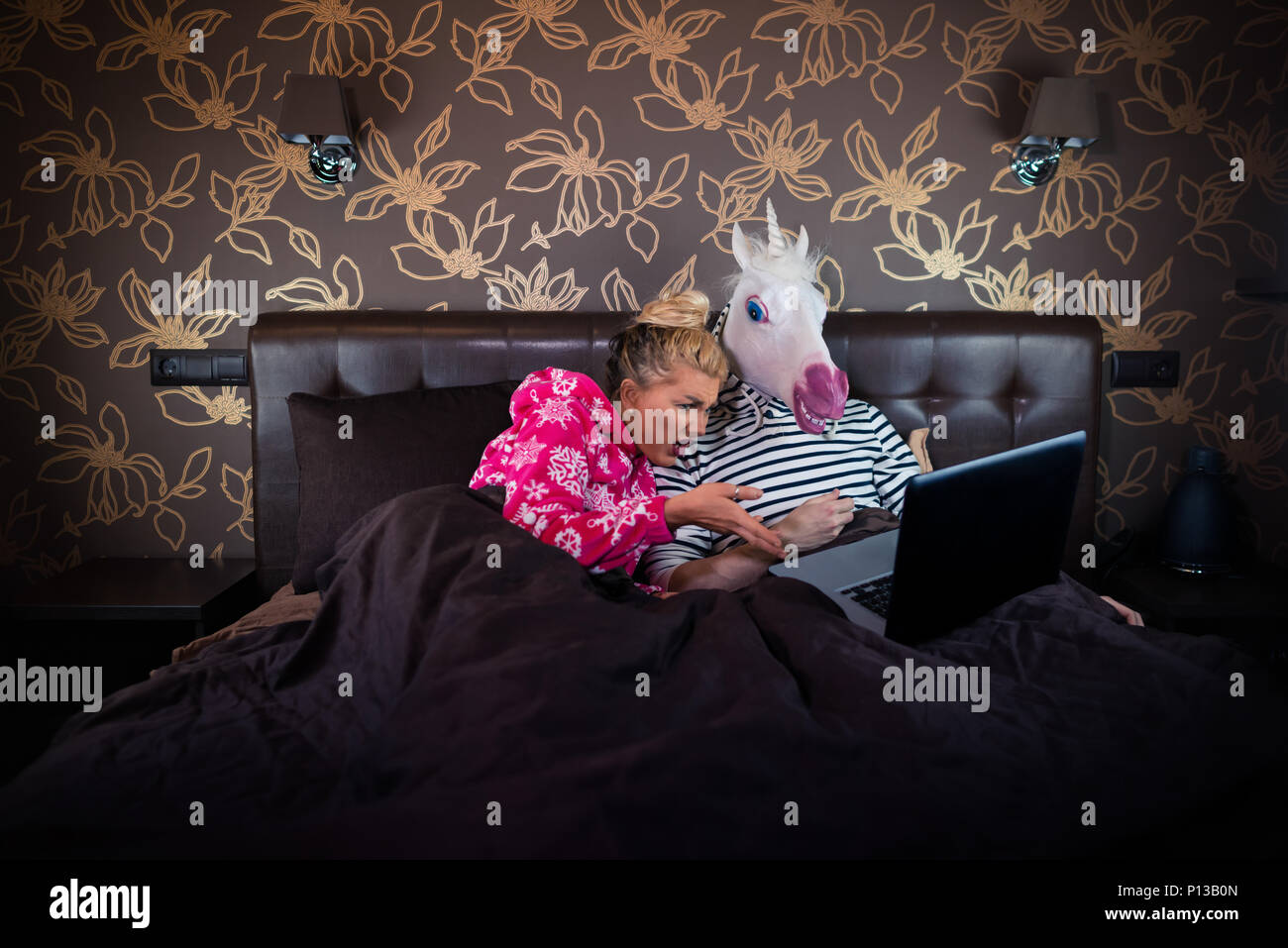 Überrascht Mädchen ist auf dem Bett liegend mit lustigen Freund in komischen Maske. Ungewöhnliche Paar beobachtet Auf der Laptop an der stilvollen Schlafzimmer. Stockfoto