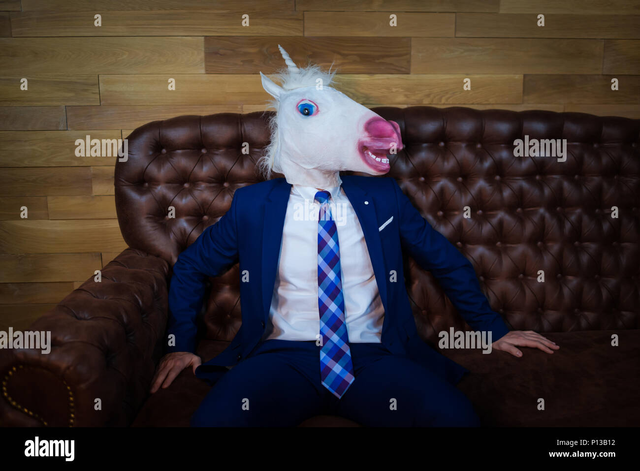 Portrait von ungewöhnlichen Menschen im Home Office. Freaky junge Manager in komischen Maske auf dem Hintergrund der hölzernen Wand. Unicorn in Anzug sitzt auf einem Sofa wie ein Chef Stockfoto