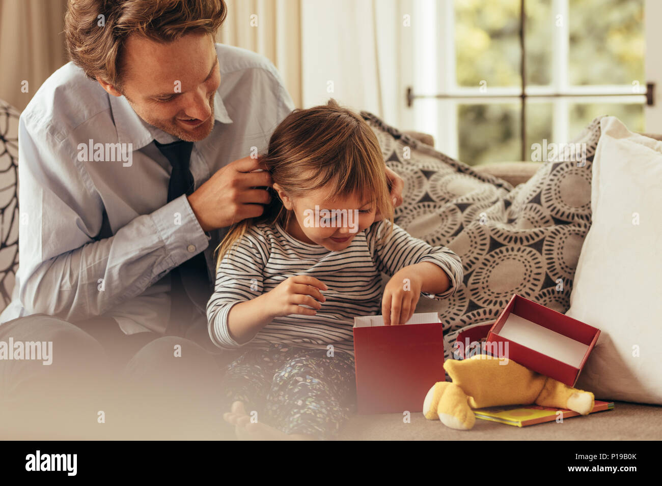 Vater und Tochter sitzen auf einem Sofa Eröffnung einer Geschenkbox. Kleines Mädchen erforscht die Geschenkbox, während ihr Vater auf aussieht. Stockfoto