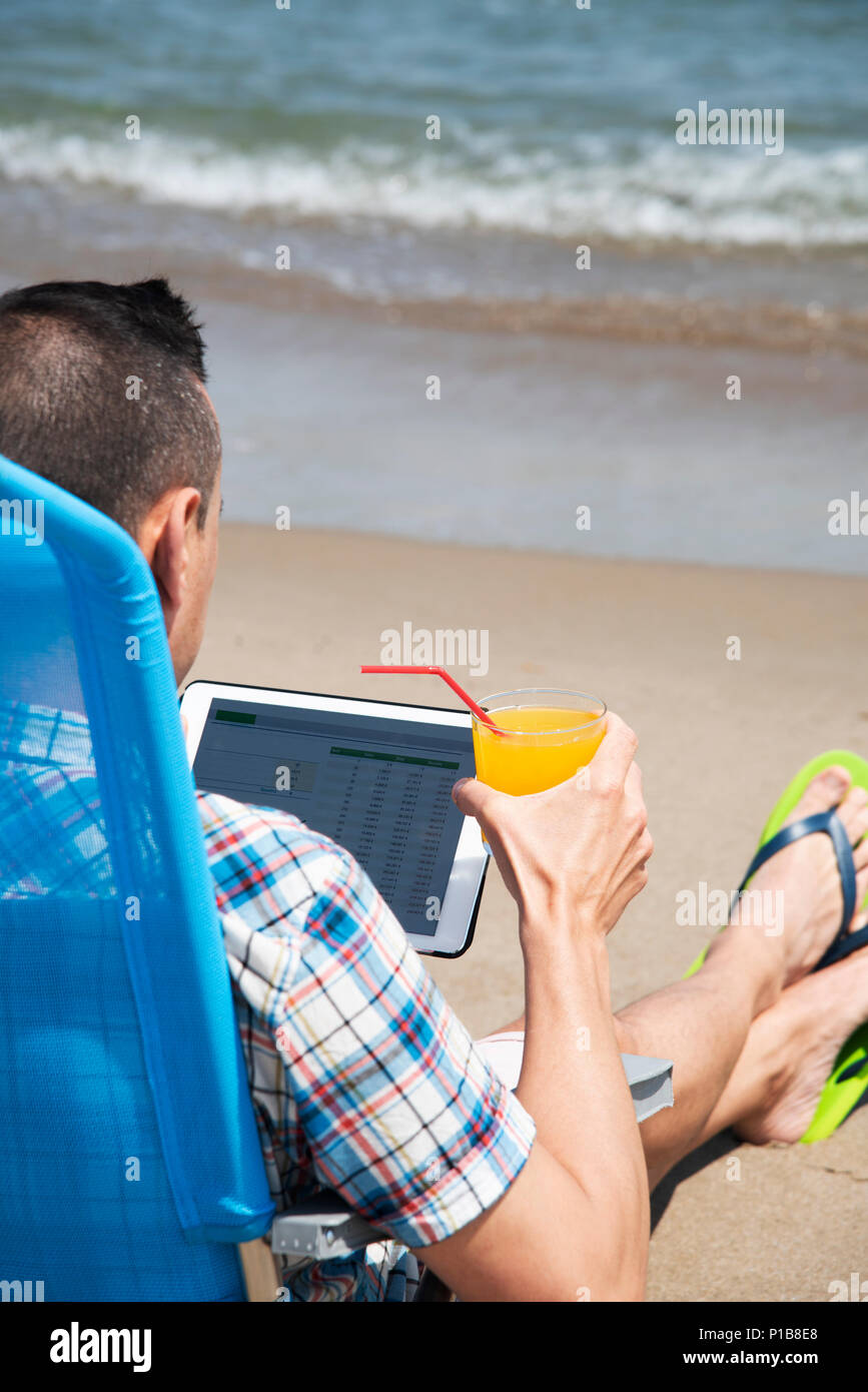 Nahaufnahme eines jungen kaukasischen Mann am Strand, mit einem digitalen Tablet, sitzend auf einem blauen Liegestuhl mit einem Glas einen erfrischenden Drink orange in seinem Stockfoto