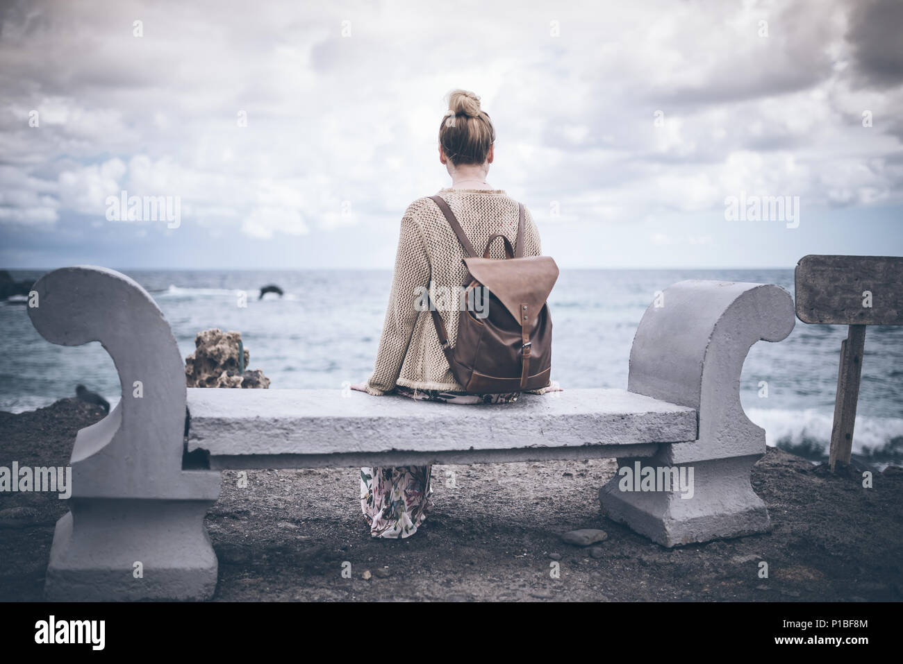Rückansicht der jungen Frau im langen Kleid und mit Rucksack sitzt auf der Bank, die auf dem Seeweg Stockfoto