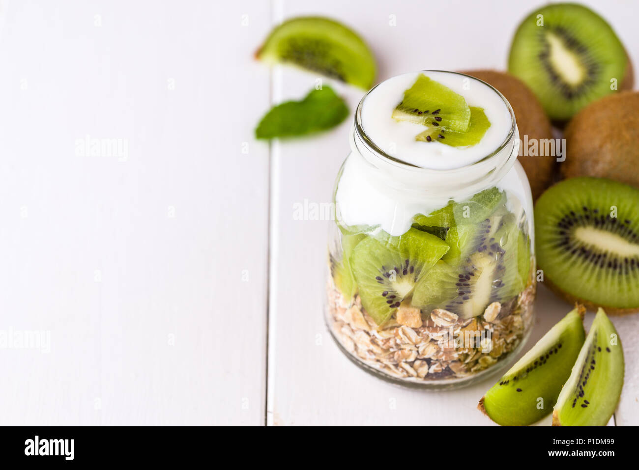 Weißer Joghurt mit Müsli in Glasschale mit Stücken von Kiwi oben und ganze Stücke auf der rechten Seite. Stockfoto