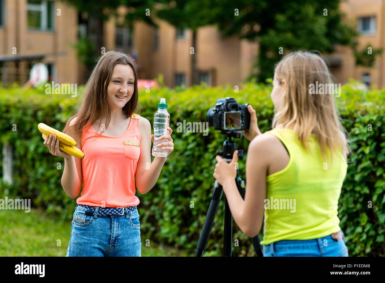 Zwei junge Mädchen. Sommer in der Natur. Erzählt über richtige Ernährung. In den Händen einer Flasche Wasser und Bananen. Vlog und blog Teilnehmer aufnehmen. Video Lektion Datensatz für das Internet. Verwenden Sie die Kamera mit einem Stativ. Stockfoto