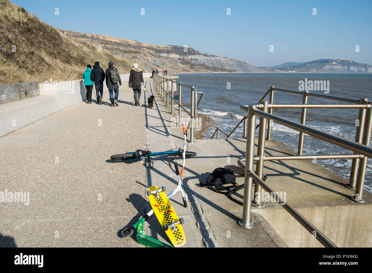 Lime Regis England Spaziergänger am Meer mit Hundeschlittenrädern Motorroller Roller mit Geländer, Pfosten und Blick auf den Küstenrand mit Seesack-Rucksack Stockfoto