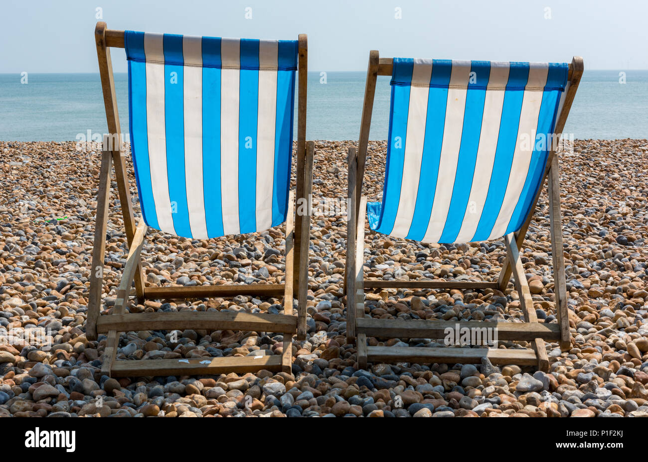 Zwei blau-weiß gestreifte oder gestreiftes altmodischen hölzernen Liegestühle leer auf dem Kiesel und Kies der Strand in Brighton, East Sussex sonnig. Stockfoto