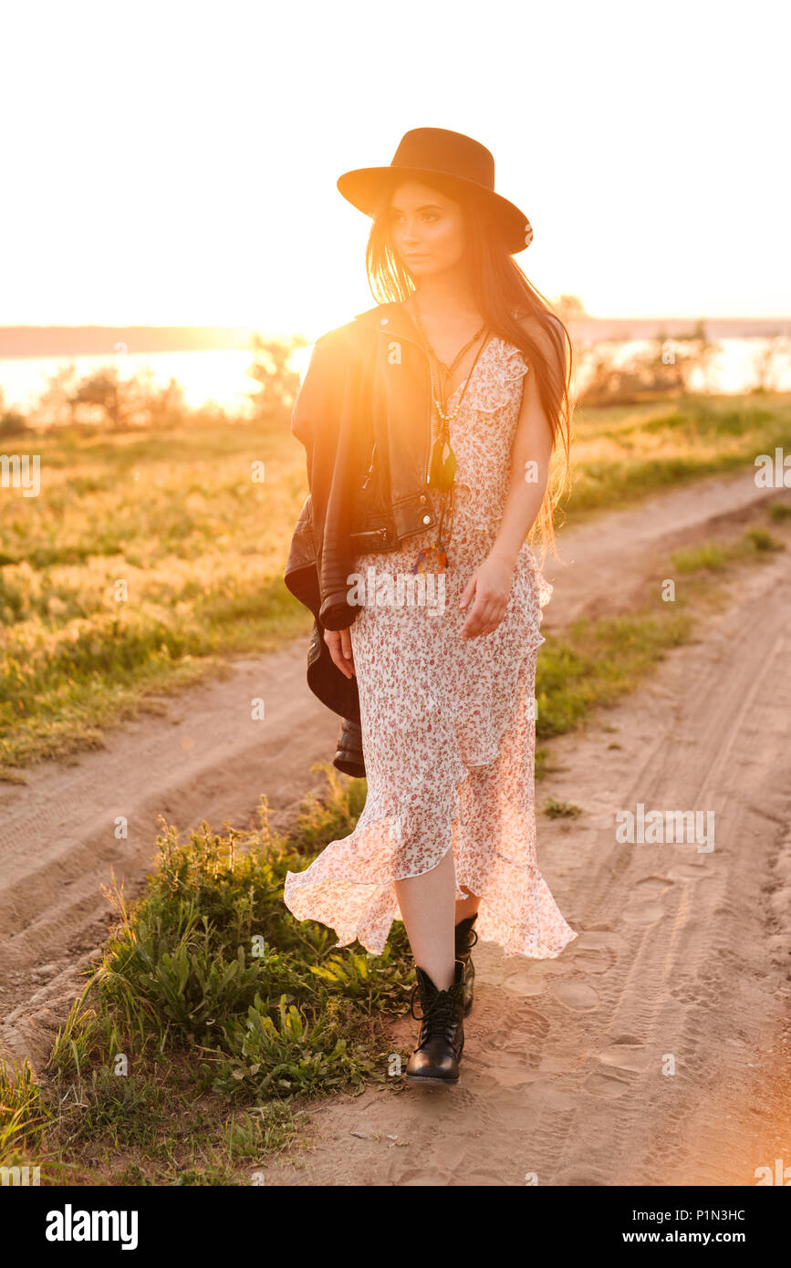 Volle Länge Bild der stilvolle kaukasische Frau mit langem Haar zu tragen kleid und hut zu Fuß auf der Straße in die Landschaft während der sonnigen warmen Tag Stockfoto