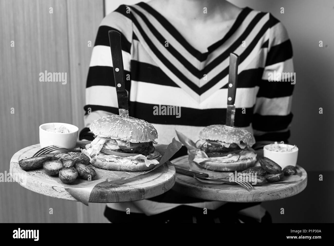 Kellnerin gekleidet in gestreiften Pullover mit Paar runden hölzernen Fächer mit leckeren Burger, Soße und Bratkartoffeln liegen auf Stücke von Kraftpapier. Stockfoto