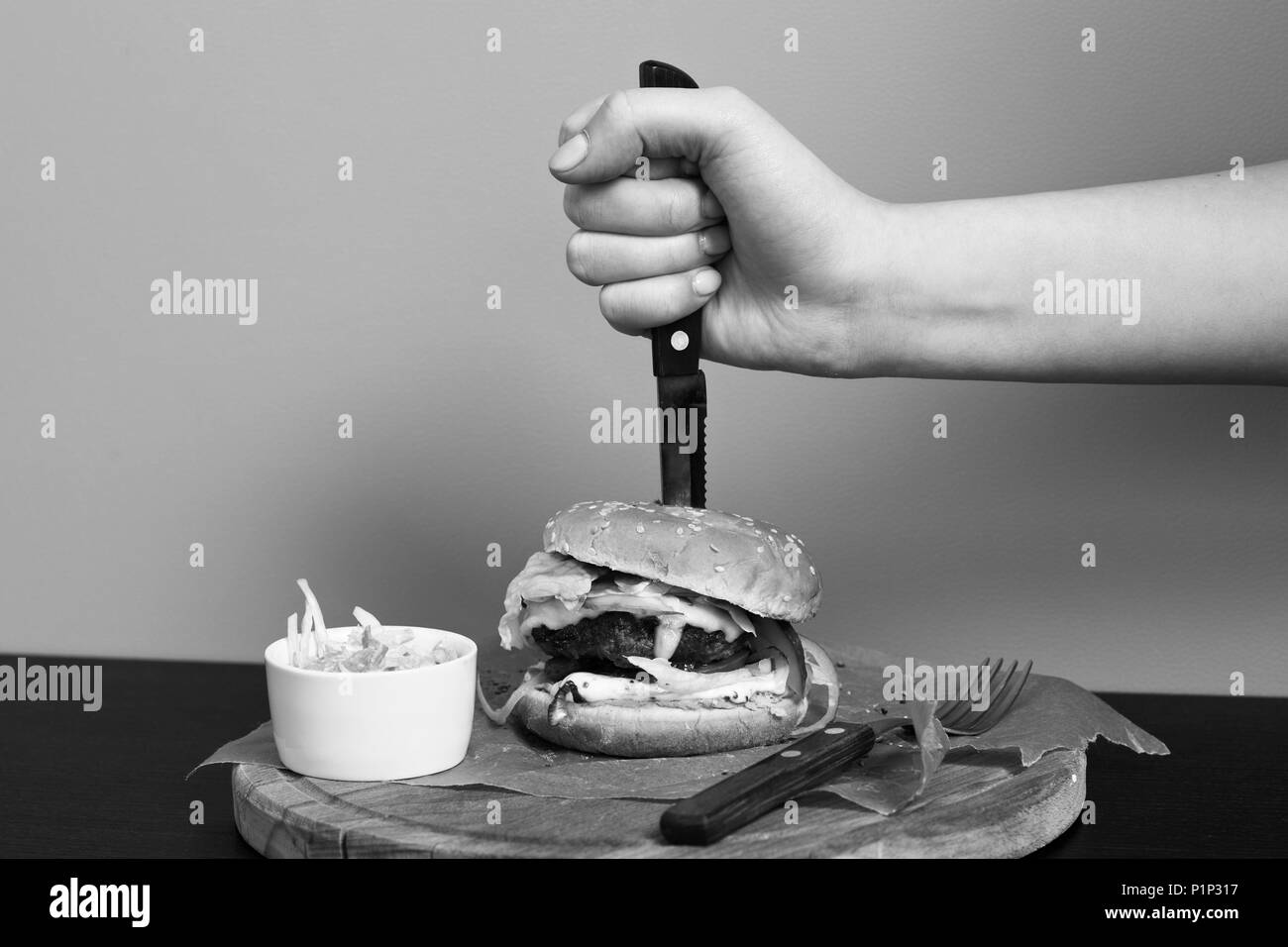 Hand mit einem Messer erstechen Burger liegen mit Portion Salat und Gabel auf runde Holzplatte mit Kraftpapier gegen orange Wand an ba abgedeckt Stockfoto