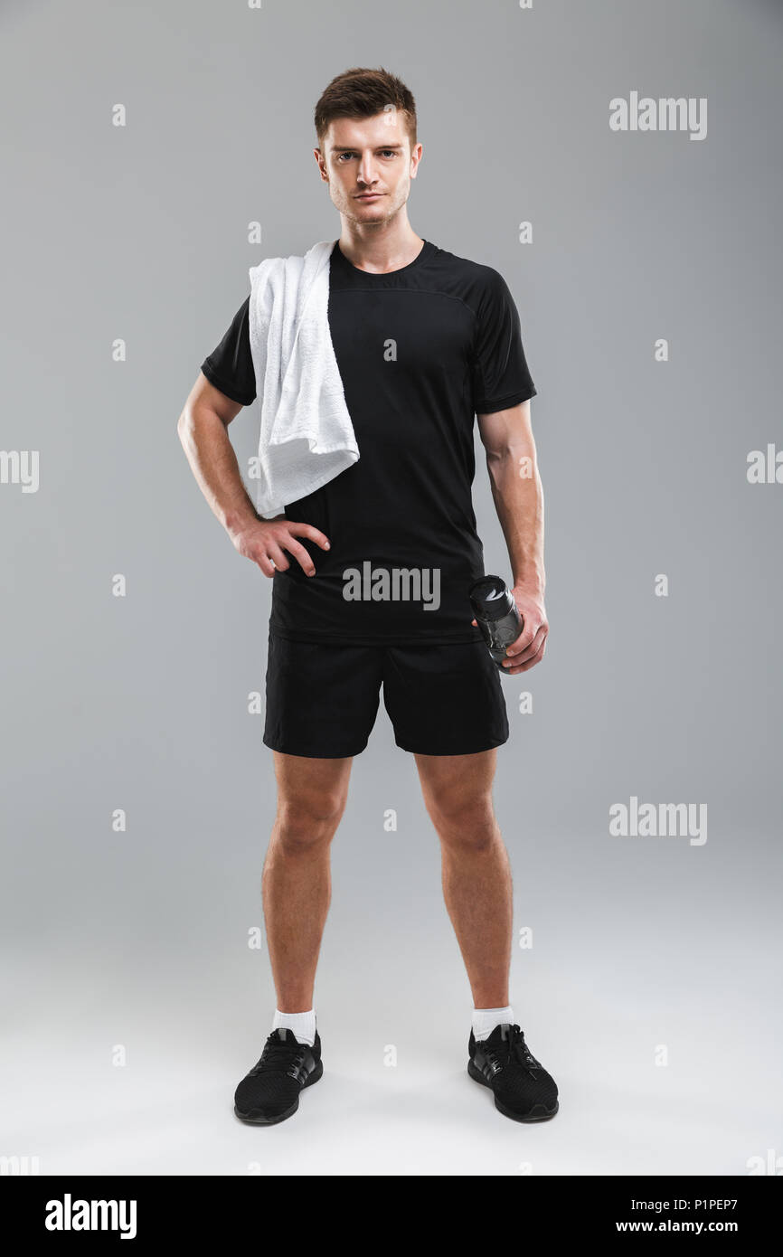 In voller Länge Porträt eines fokussierten junge Sportler mit Wasserflasche und Handtuch auf seiner Schulter isoliert über grauer Hintergrund Stockfoto