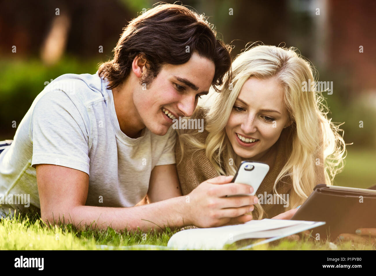 Ein junges Paar legt Studieren im Freien auf dem Gras auf dem Campus der Universität mit einem Lehrbuch und Tablet und die Überprüfung der sozialen Medien auf einem Smartphone Stockfoto