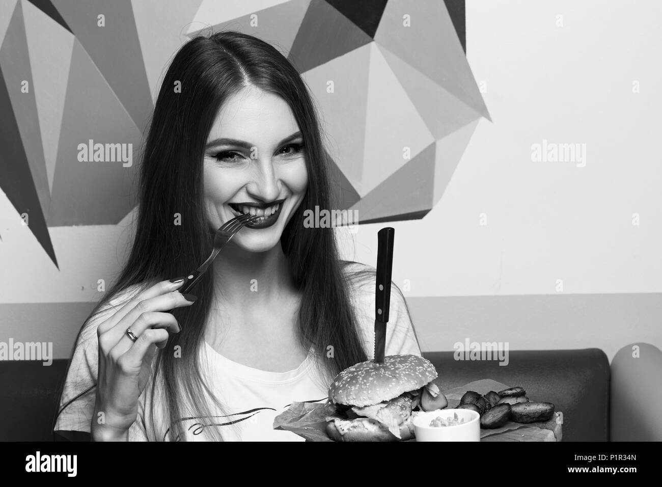 Gerne attraktive Frau mit Gabel und runde Holzplatte mit appetitlichen Burger und gebratenen Kartoffeln. Schöne fröhliche weibliche Modell essen Cheeseburger. Stockfoto