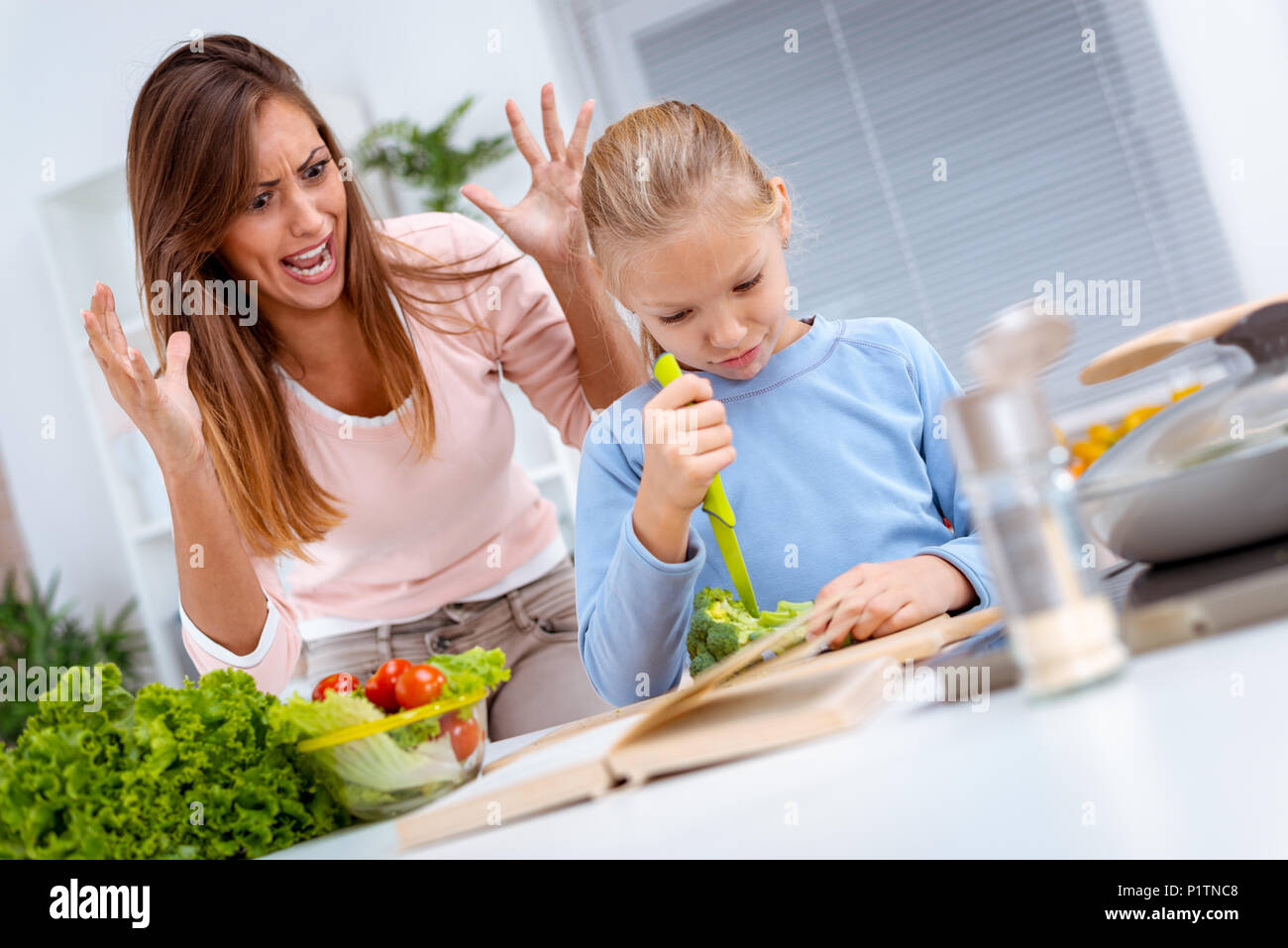 Ein kleines Mädchen versucht, Brokkoli allein zu schneiden und ihre Mutter macht sich Sorgen, dass Sie sich verletzt. Stockfoto