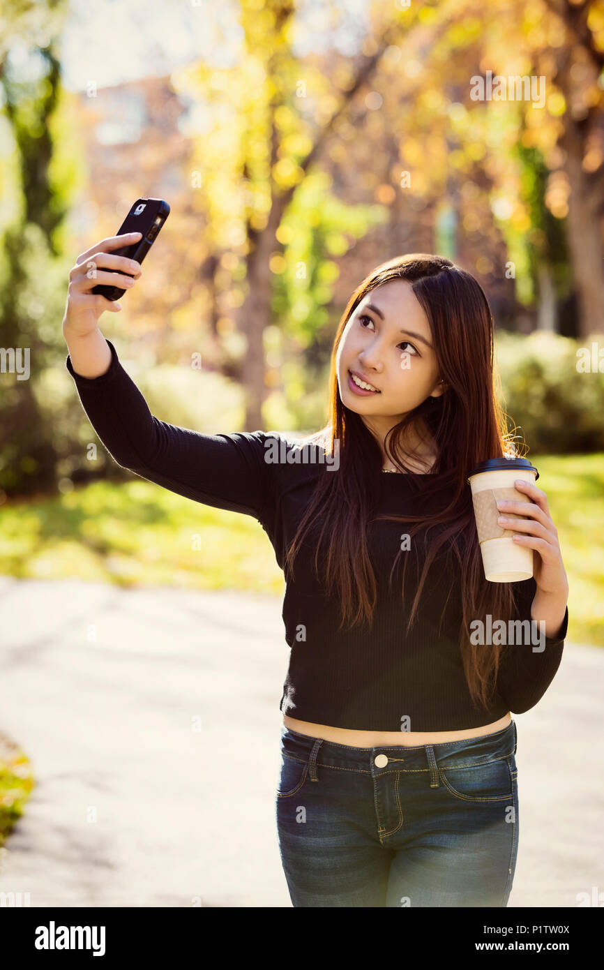 Eine junge weibliche International University Student steht eine Kaffeetasse und posiert für ein Selbstporträt mit Ihrem Smart Phone Stockfoto