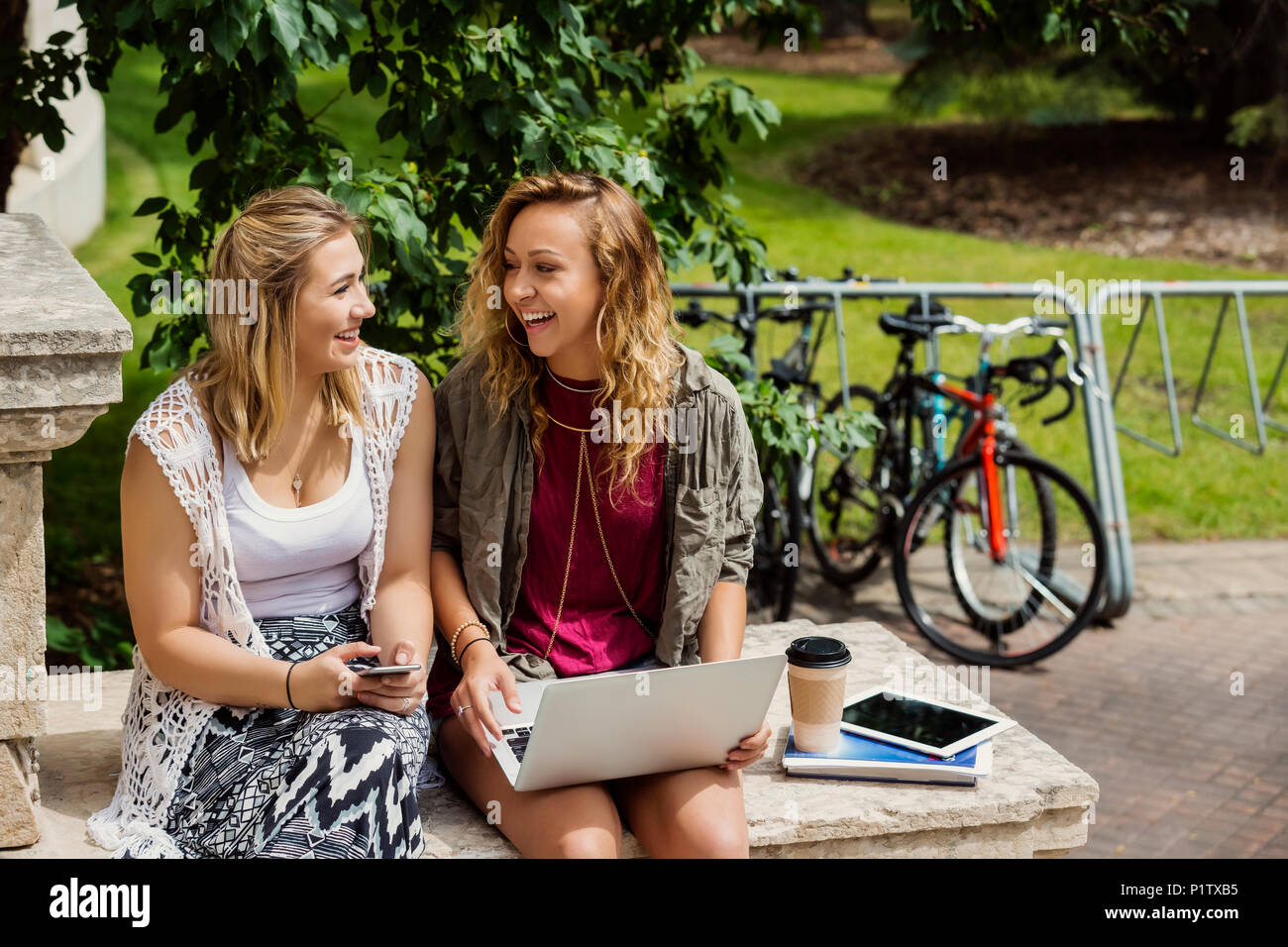 Zwei weibliche Studenten lachen und miteinander sprechen, während Sie Ihre Technologie außerhalb auf dem Campus; Edmonton, Alberta, Kanada Stockfoto