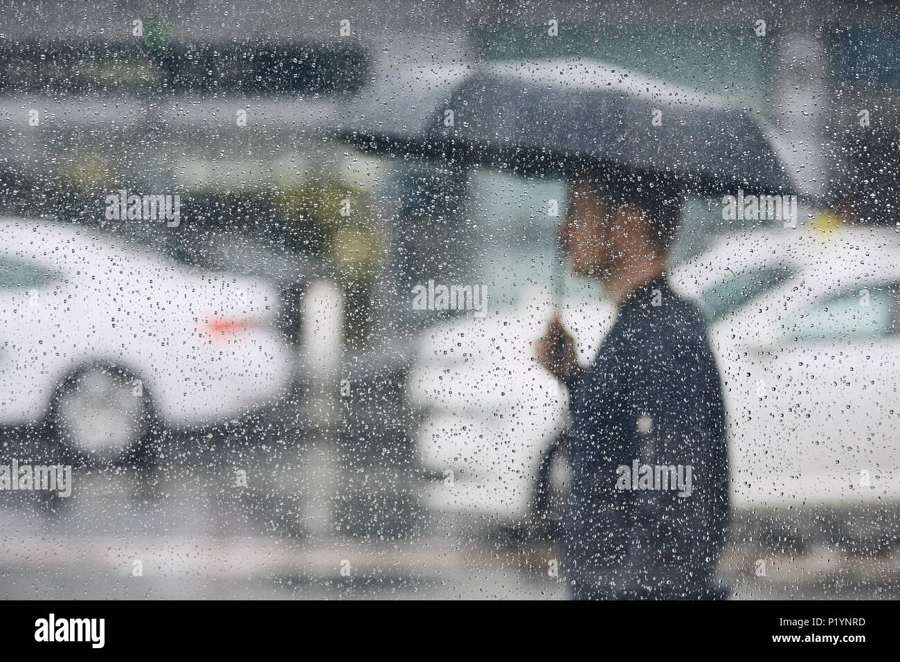 Regen in der Stadt. Junger Mann mit Regenschirm zu Fuß auf der Straße. Selektiver Fokus auf Regentropfen auf dem Fenster. Stockfoto