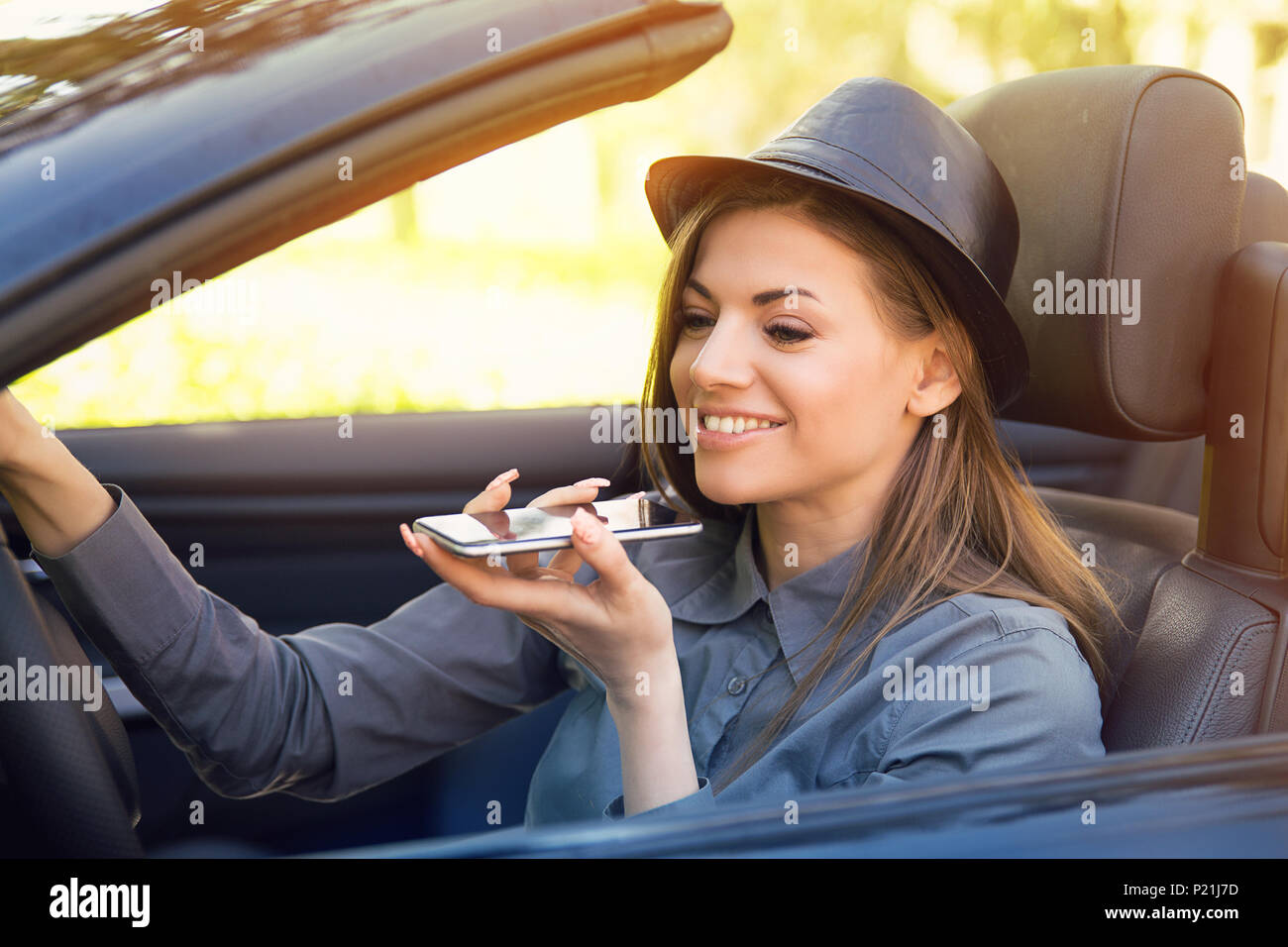 Gerne Frau sitzt im Inneren Cabrio mit einem smart phone Funktion Spracherkennung auf der Linie im Freien auf einem beleuchteten Park Hintergrund Stockfoto