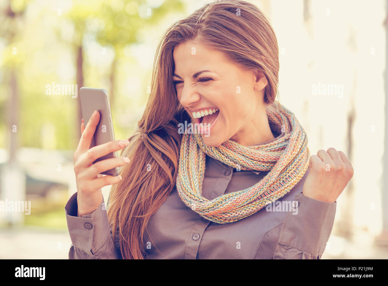 Begeistert Schüler lesen gute Nachrichten auf Handy im Freien an einem warmen Herbsttag mit sonnendurchfluteten Straße Hintergrund Stockfoto