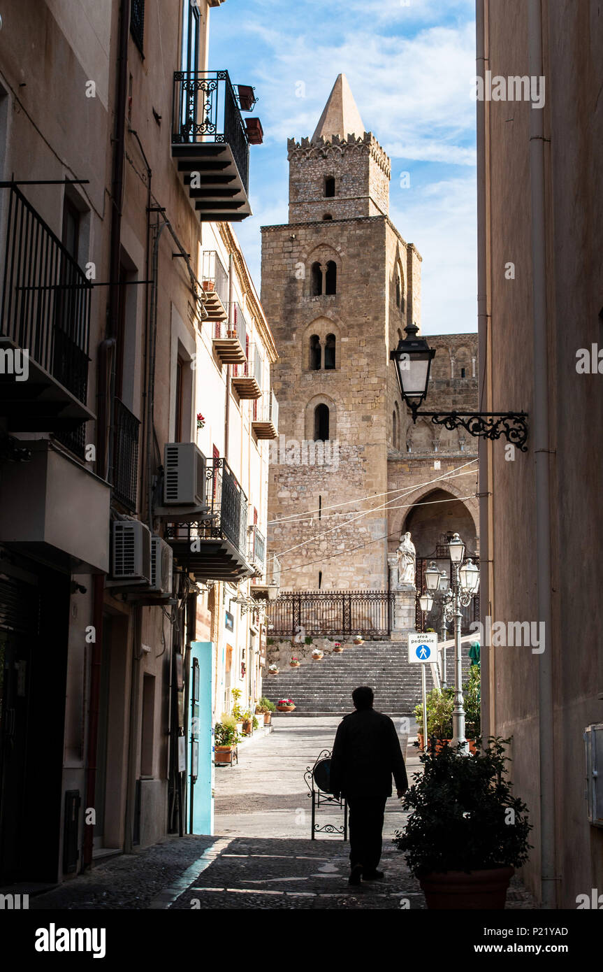 Ein Mann wird man über eine kleine Straße in der mittelalterlichen Altstadt von Cefalu. Vor ihm, die beeindruckende normannische Kathedrale der Stadt. Sizilien, Italien. Stockfoto