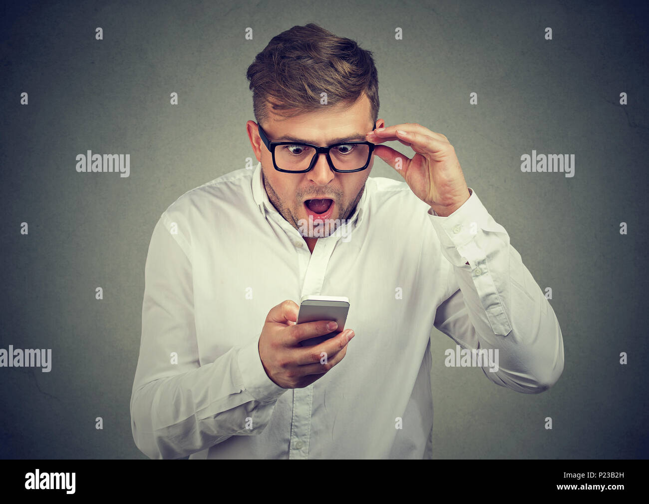 Mann in T-Shirt und Brillen mit Nachrichten erstaunt beim Surfen Smartphone auf grauem Hintergrund. Stockfoto