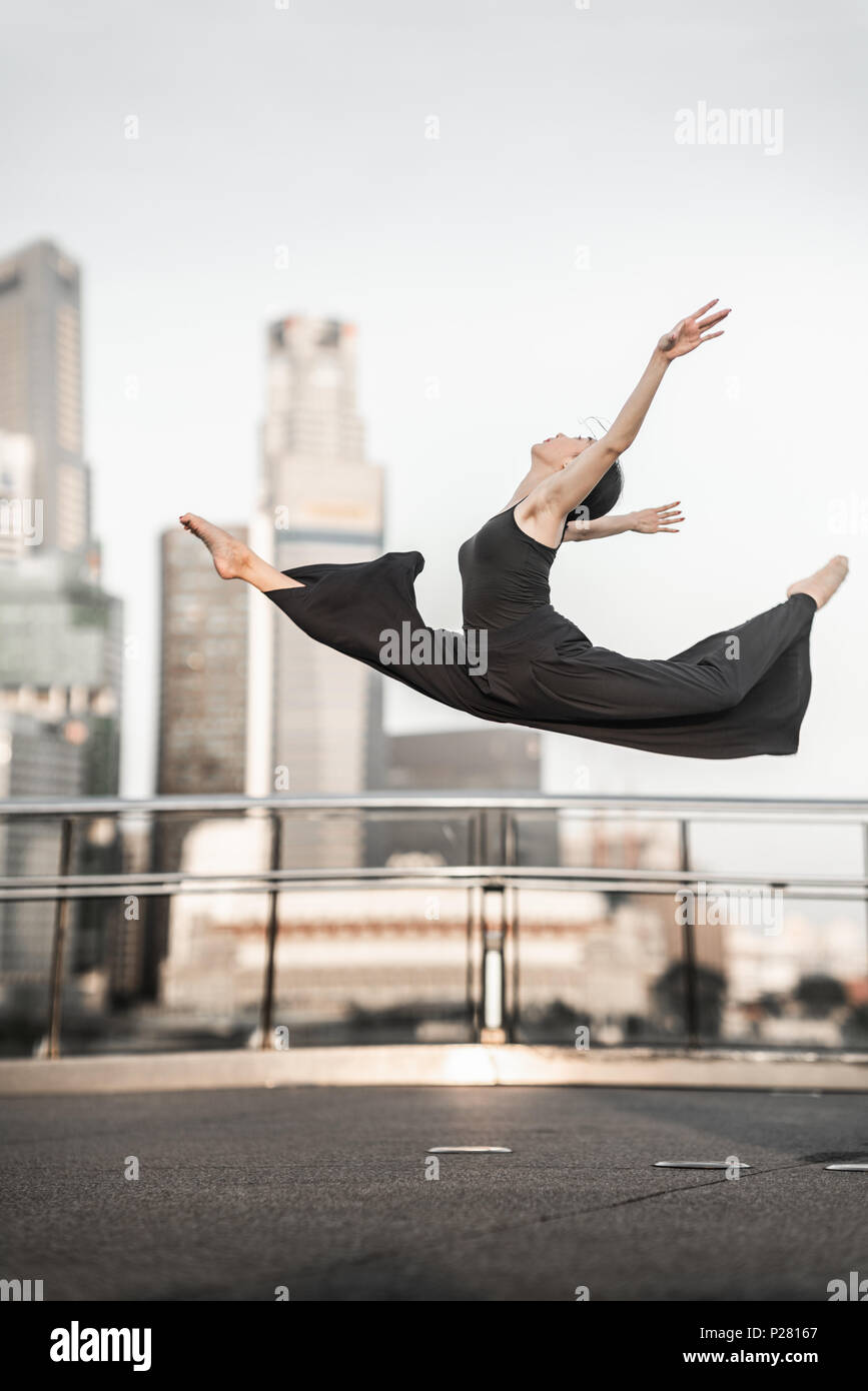 Nette junge Sportlerin führt eine perfekte straddle Sprung hoch, mit perfekter Kontrolle beim Lächeln, auf einer Brücke mit Hintergrund der Wolkenkratzer Stockfoto