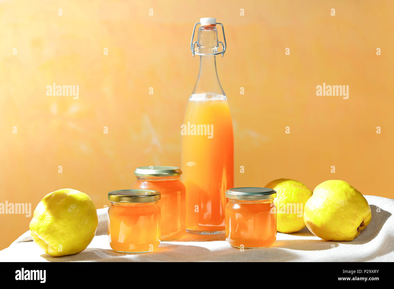 Hausgemachte Quitten Gelee und Saft in Flaschen und Gläsern mit Quitten auf Leinen Tischdecke im hellen Sonnenschein vor einem gelben und orangefarbenen Hintergrund Stockfoto